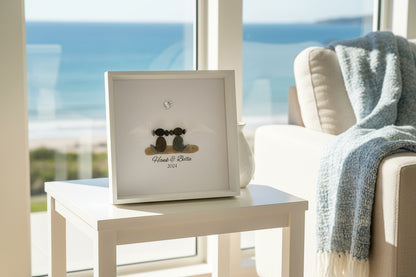 A personalized dog memorial gift featuring a sea glass and pebble art with the names 'Hank & Bella' and a date '2024' inscribed on a piece of paper within a black frame.