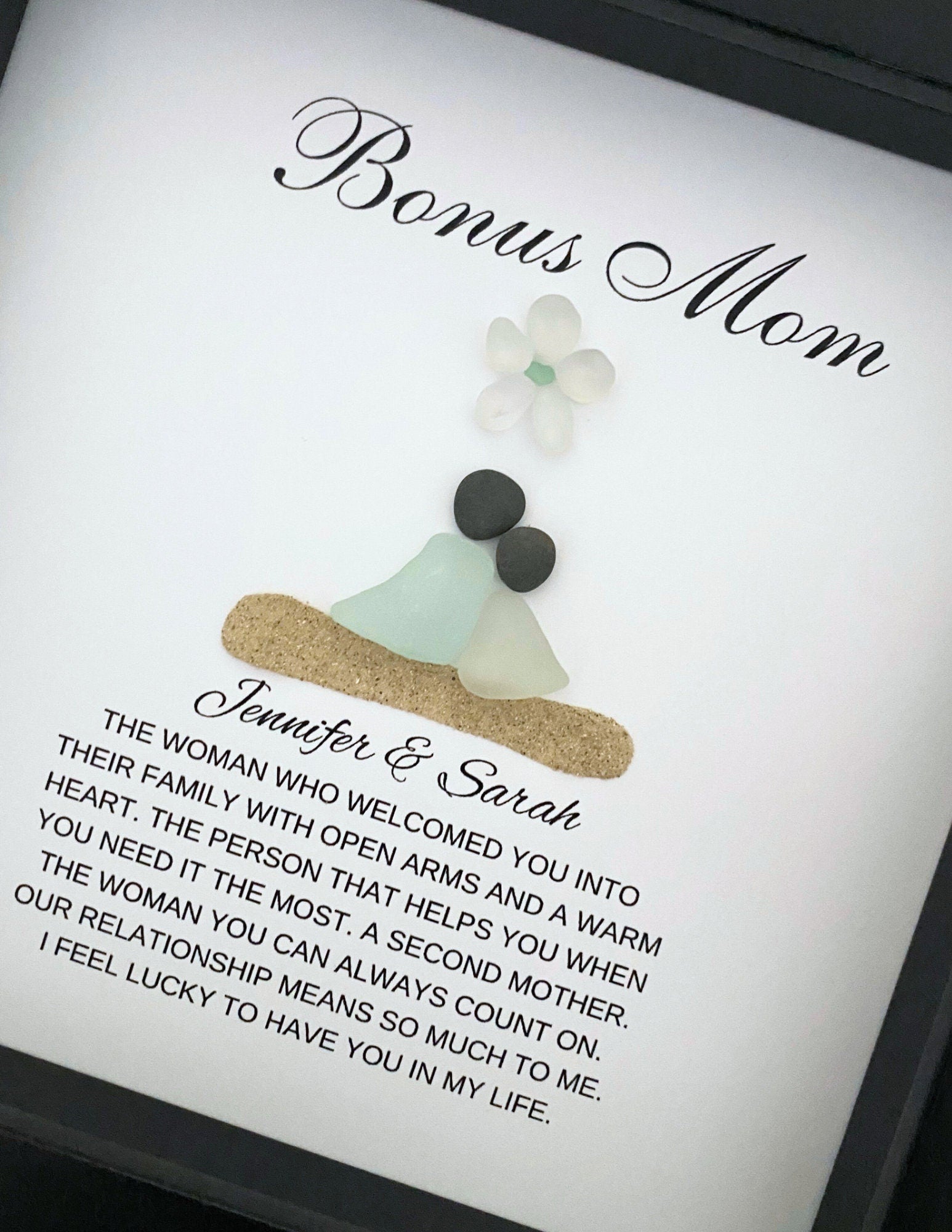 A personalized gift frame with a title 'BONUS MOM' at the top, featuring a sea glass figure and pebble art, placed on a soft surface.