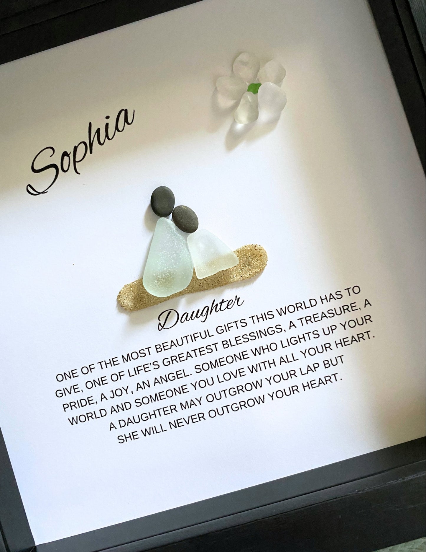 A framed pebble art piece with sea glass and stones, personalized with the name 'Sophia' and a message from a mother to her daughter.