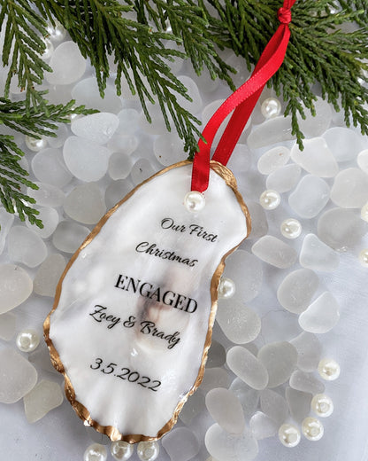 A personalized engagement ornament made of oyster shell with gold gilded edges, displaying a message that reads 'Our First Christmas Engaged', with a date '3.2.2022' below the text. The ornament is hanging from a red ribbon, placed against a backdrop of pine needles.