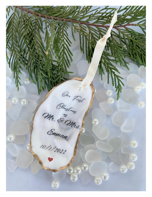 A personalized oyster shell Christmas ornament with the text 'Mr & Mrs' and a date '10/1/2023' on it, hanging from a branch with pearl-like beads and greenery in the background.