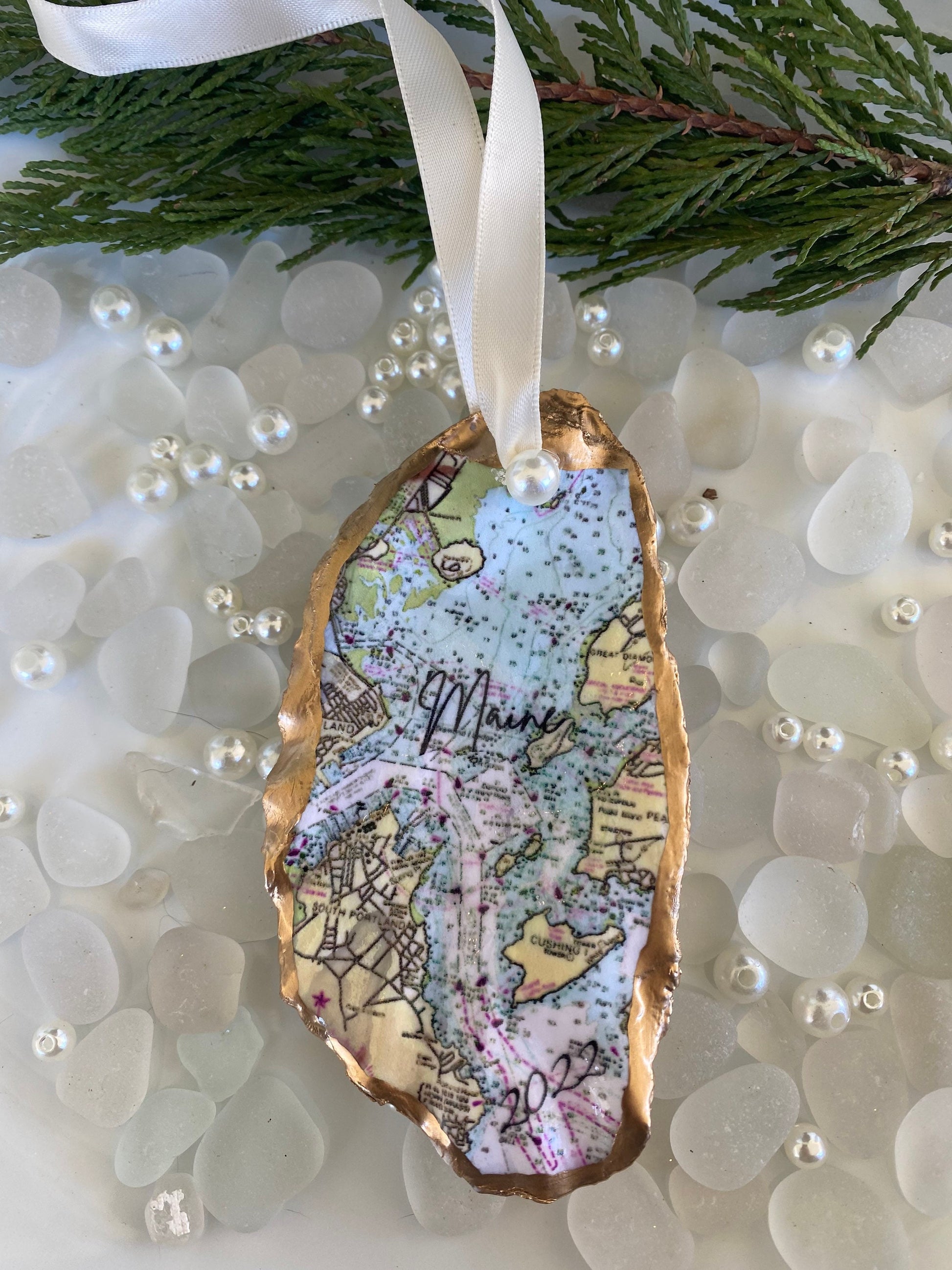 A custom Casco Bay Maine map oyster shell Christmas ornament with the word 'Maine' written on it, personalized with a name and date, displayed on a white background with pearls and greenery.