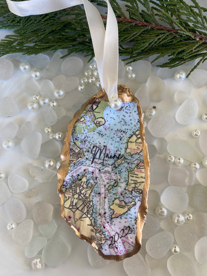 A custom Casco Bay Maine map oyster shell Christmas ornament with the word 'Maine' written on it, personalized with a name and date, displayed on a white background with pearls and greenery.