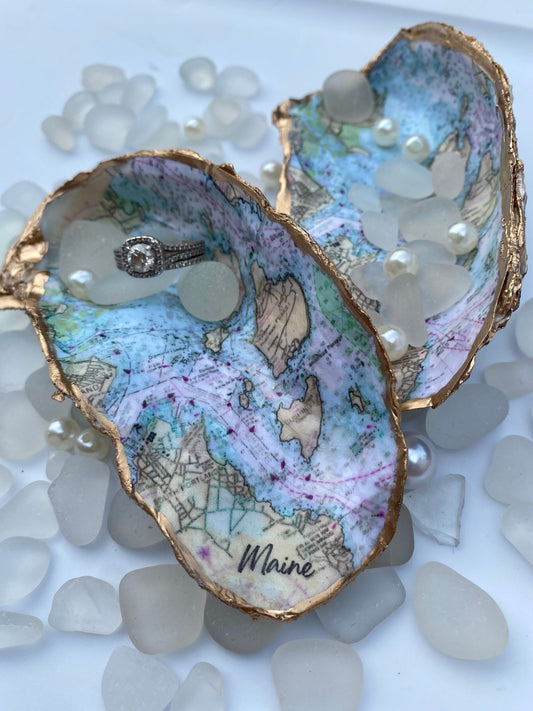A custom map oyster dish featuring a hand-painted design of the state of Maine, with inlays of pearls and a ring, displayed on a white surface with additional pearls scattered around.