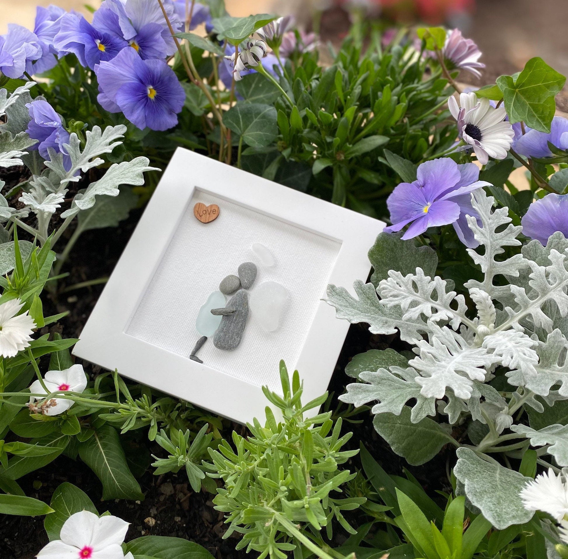 A framed sea glass art piece with a white frame, featuring a picture of a mother embracing her child angel with a wooden heart.