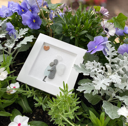 A framed sea glass art piece with a white frame, featuring a picture of a mother embracing her child angel with a wooden heart.