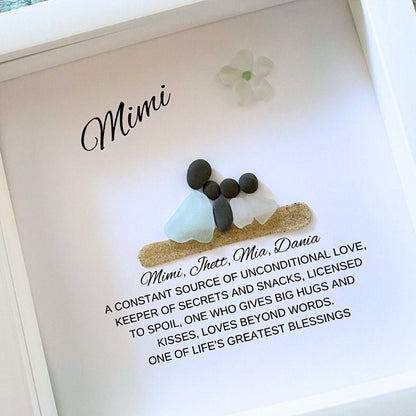 A personalized sea glass and pebble art piece with the inscription 'Mimi' in a shadowbox frame, accompanied by a floral decoration.