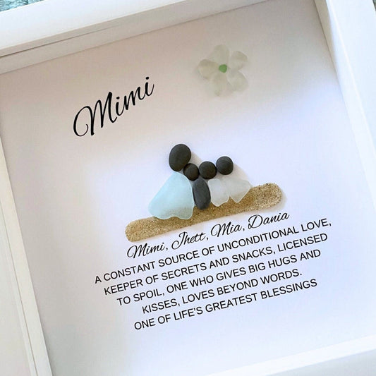 A personalized sea glass and pebble art piece with the inscription 'Mimi' in a shadowbox frame, accompanied by a floral decoration.