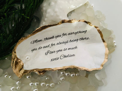 A personalized oyster shell jewelry dish with a message written on it, placed on a white surface surrounded by decorative pearls.