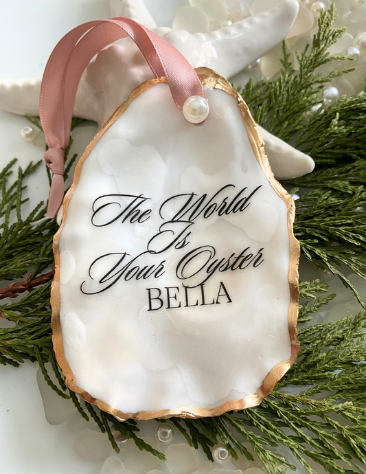 A personalized oyster shell ornament with the inscription 'The World is Your Oyster' and a name 'Bella' on it, with a gold gilded edge, displayed on a branch with green leaves and pearls.