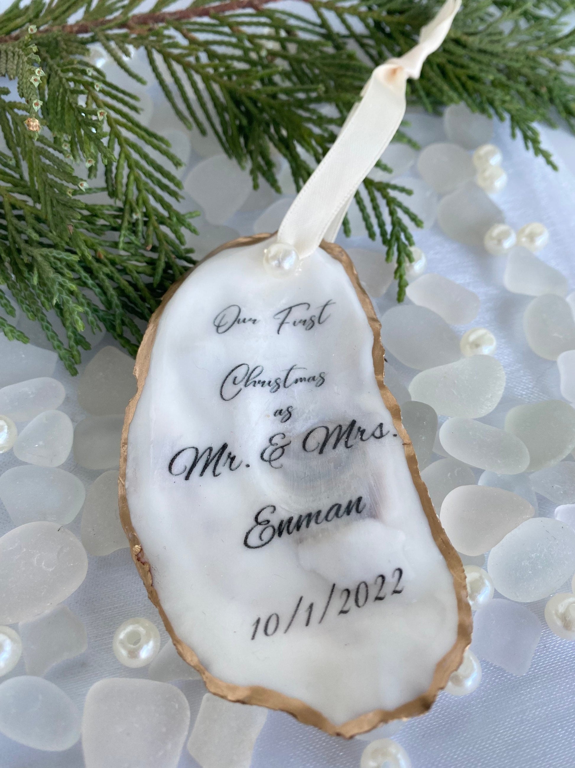 A personalized wedding Christmas ornament made of oyster shell with the names 'Mr & Mrs' and a date '11/27/2024'  on it, placed on a branch with pine needles and decorative pearls around.