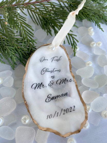A personalized wedding Christmas ornament made of oyster shell with the names 'Mr & Mrs' and a date '11/27/2024'  on it, placed on a branch with pine needles and decorative pearls around.