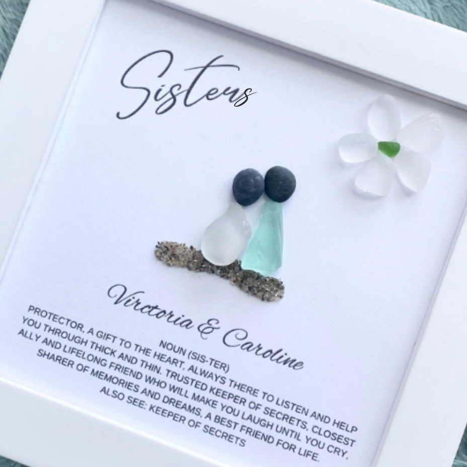 A framed personalized art piece with the word 'Sisters' at the top, featuring sea glass and pebble art, placed on a soft surface.