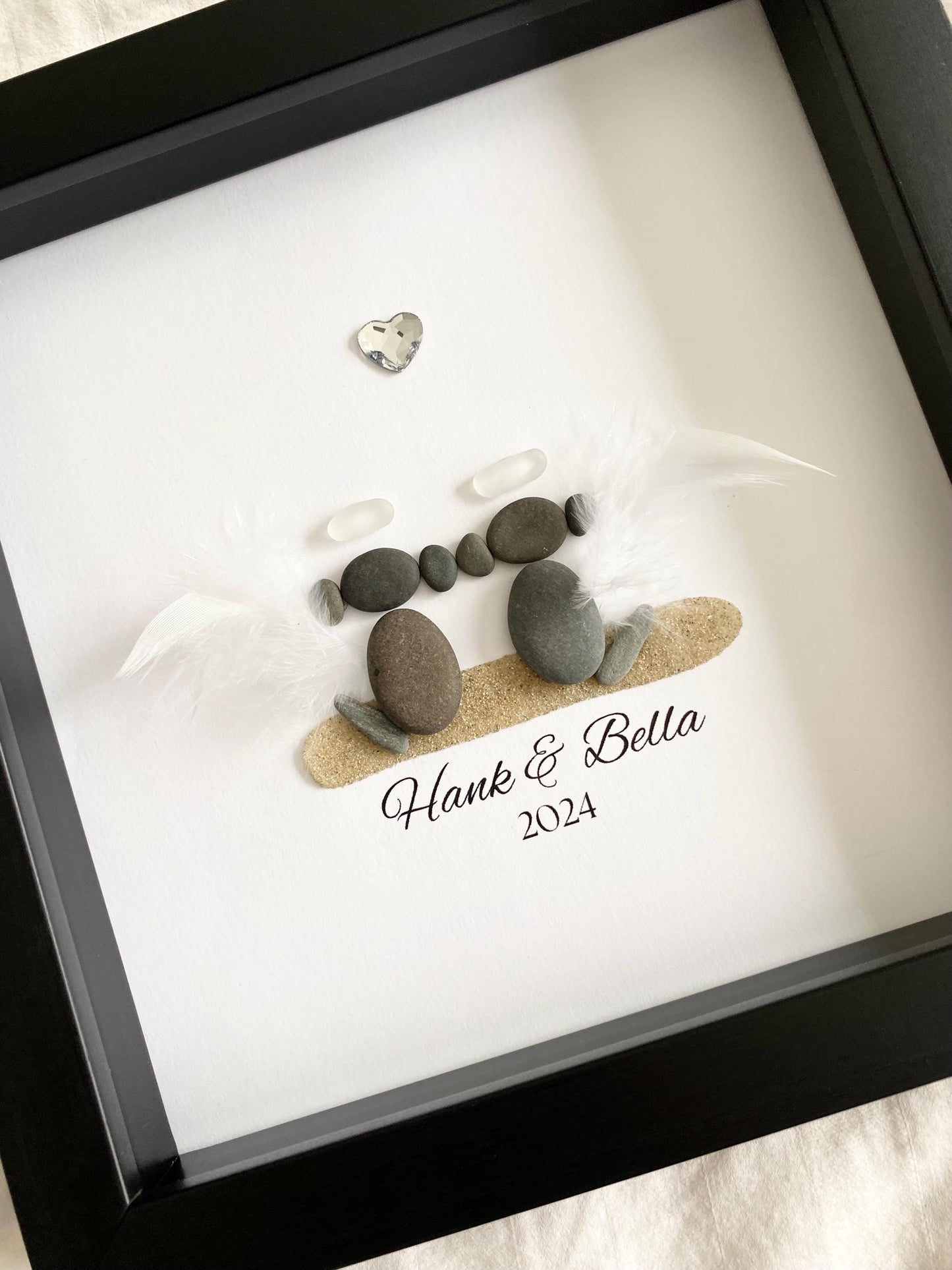 A personalized dog memorial gift featuring a sea glass and pebble art with the names 'Hank & Bella' and a date '2024' inscribed on a piece of paper within a black frame.