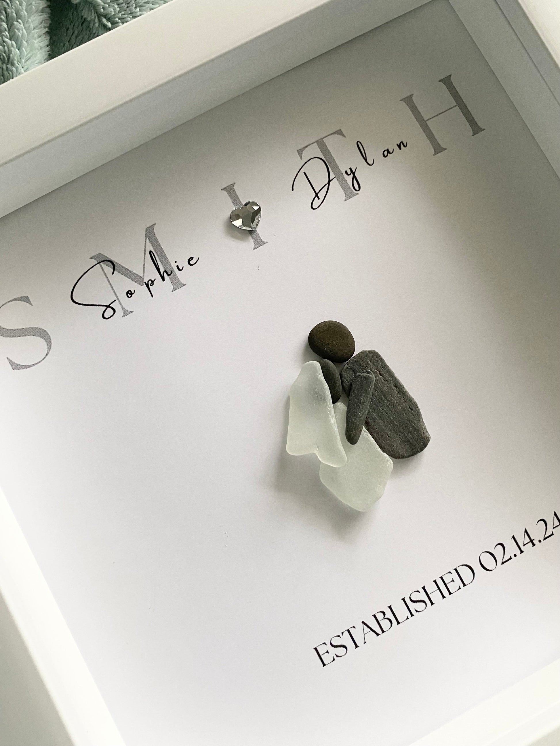A framed personalized sea glass art piece with the names 'Sophie & Ryan' and a date 'September 7, 2022' written on it, displayed in a white frame.