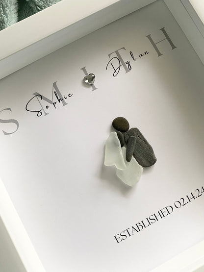 A framed personalized sea glass art piece with the names 'Sophie & Ryan' and a date 'September 7, 2022' written on it, displayed in a white frame.