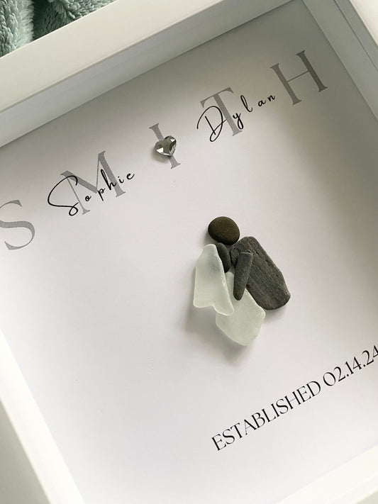 A framed personalized sea glass art piece with the names 'Sophie & Ryan' and a date 'September 7, 2022' written on it, displayed in a white frame.