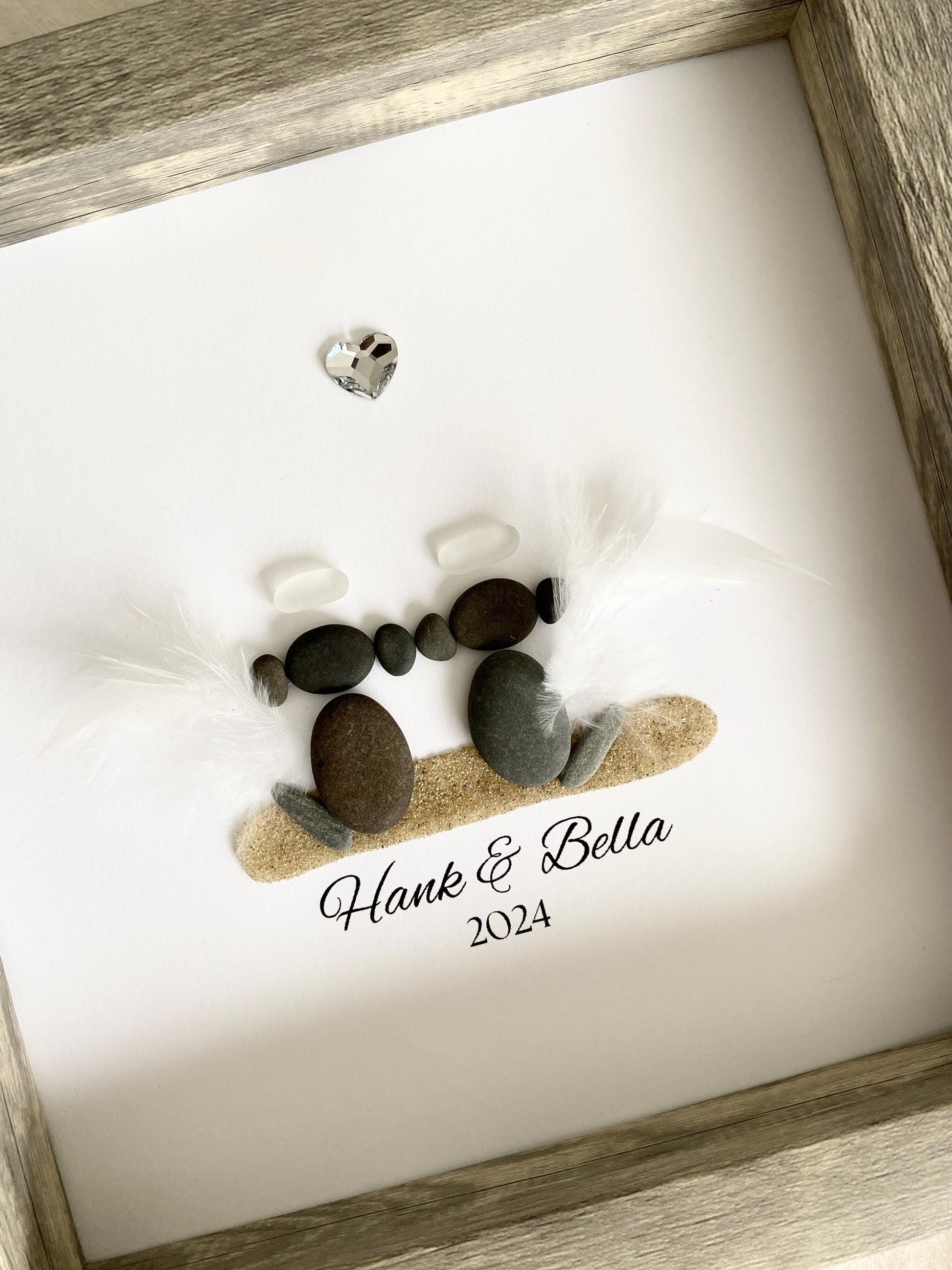 A personalized dog memorial gift featuring a sea glass and pebble art with the names 'Hank & Bella' and a date '2024' inscribed on a piece of paper within a gray frame.