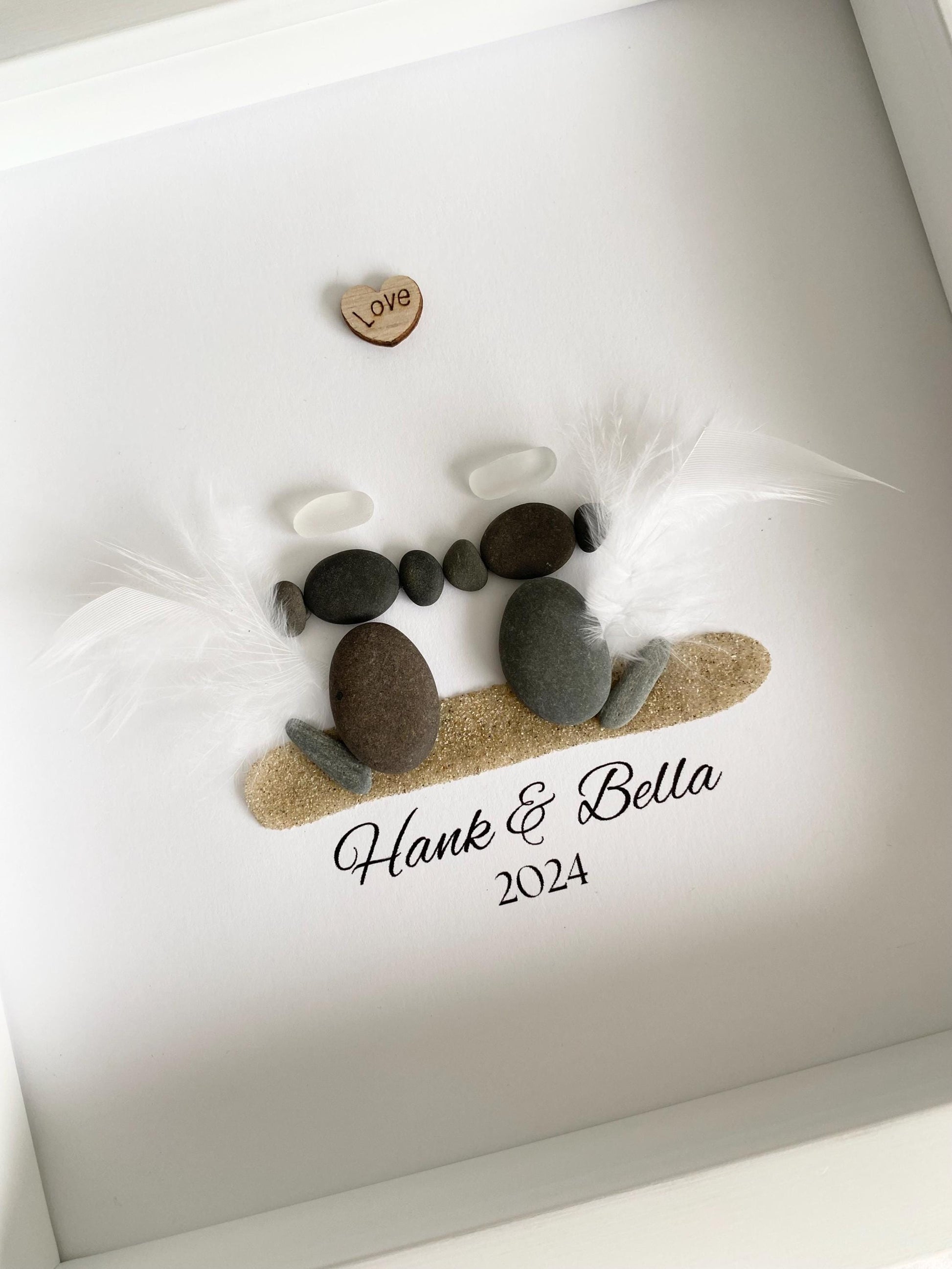 A personalized dog memorial gift featuring a sea glass and pebble art with the names 'Hank & Bella' and a date '2024' inscribed on a piece of paper within a white frame.