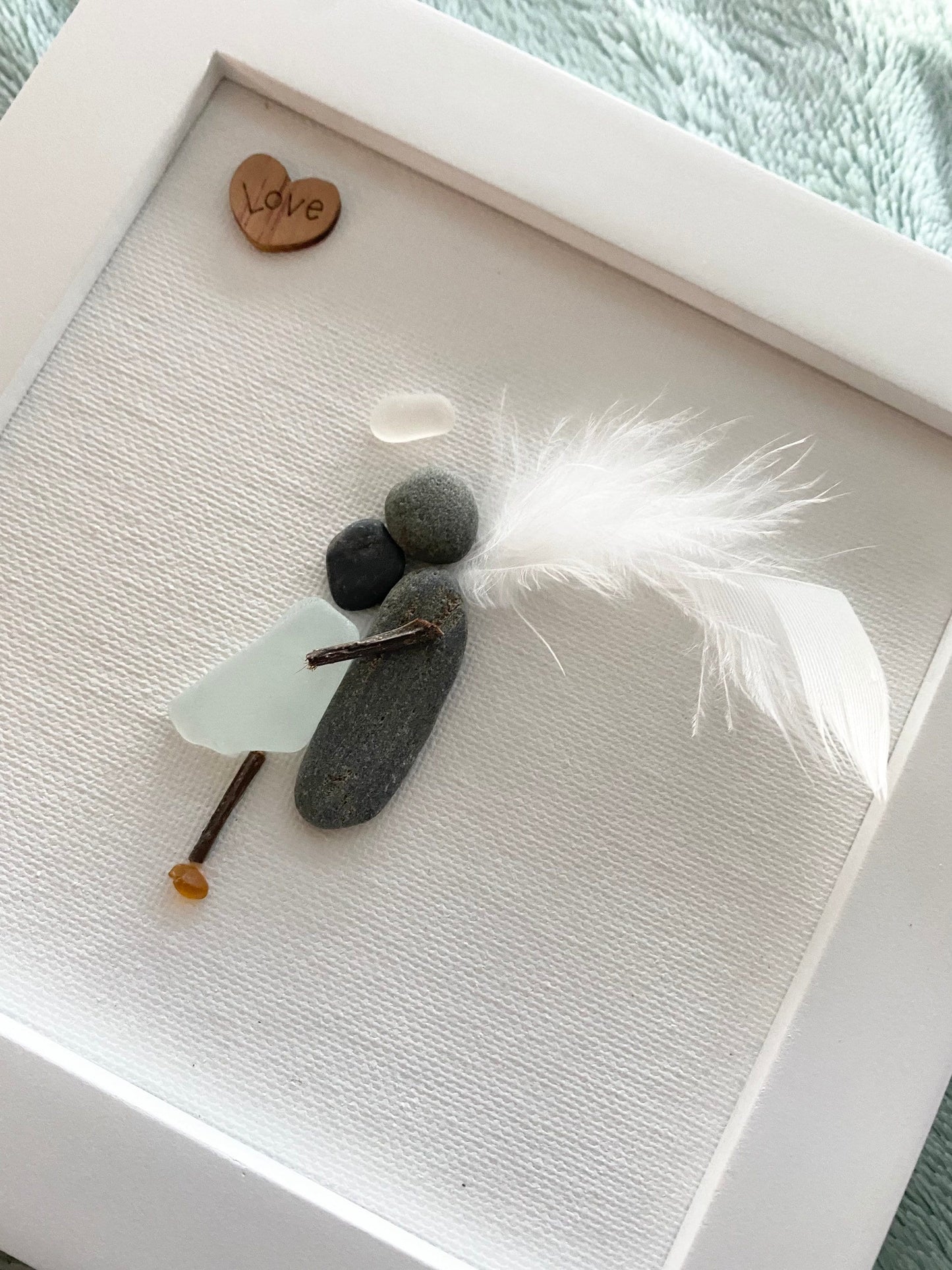A framed sea glass art piece with a white frame, featuring a picture of a mother embracing her child angel with a wooden heart.