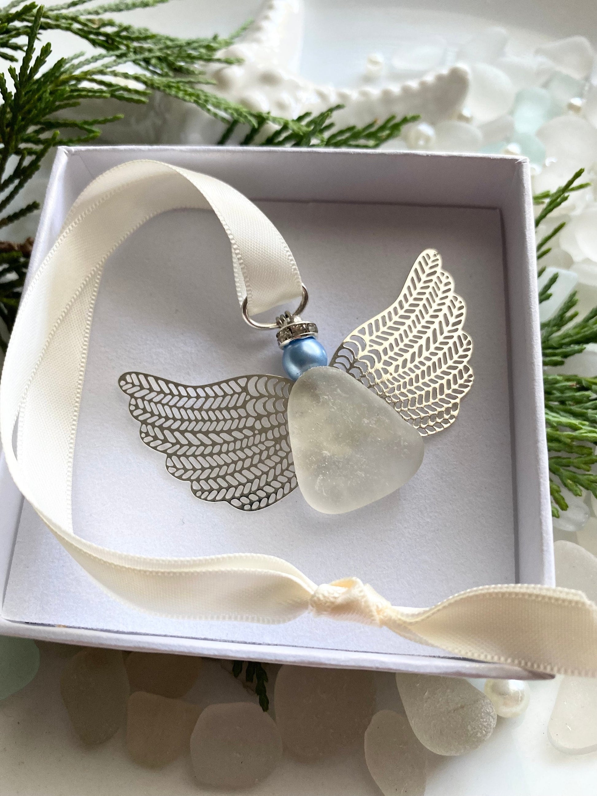 A silver-colored angel-shaped ornament made of sea glass with a blue pearl head, displayed in an open white box with a satin ribbon, set against a natural background with sea glass accents.
