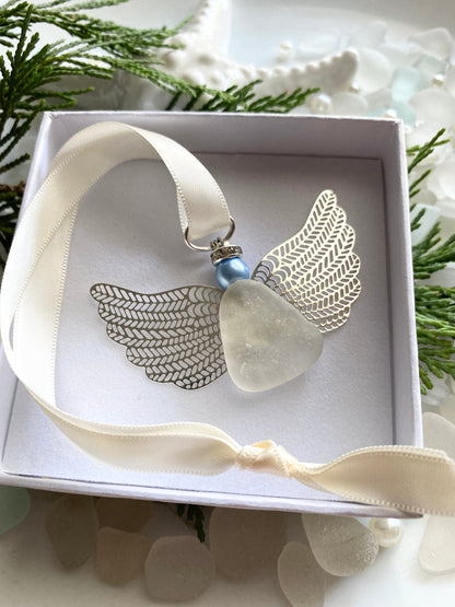 A silver-colored angel-shaped ornament made of sea glass with a blue pearl head, displayed in an open white box with a satin ribbon, set against a natural background with sea glass accents.