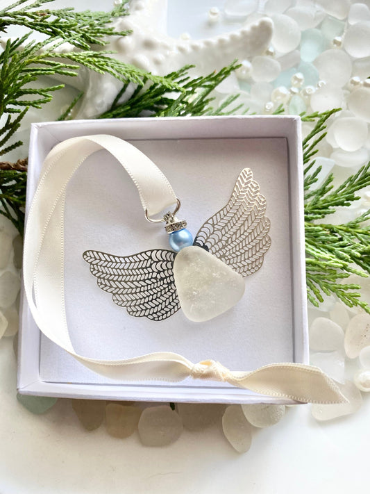 A silver-colored angel-shaped ornament made of sea glass with a blue pearl head, displayed in an open white box with a satin ribbon, set against a natural background with sea glass accents.