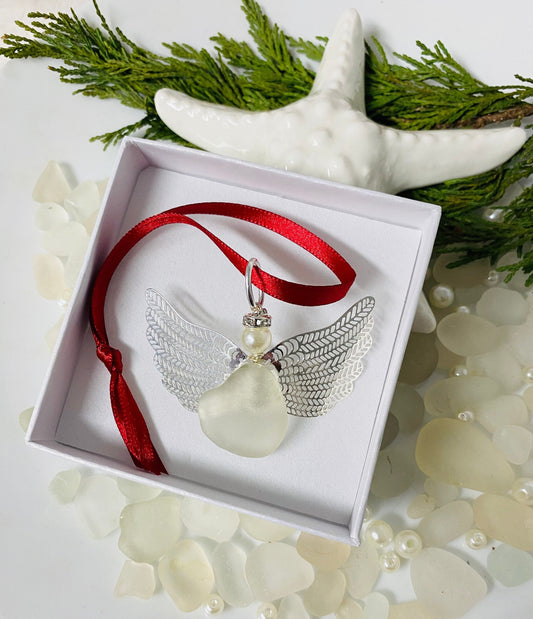 A handcrafted sea glass angel Christmas ornament with a red ribbon, displayed in a white box with a white starfish and pine needles around it, on a white background.