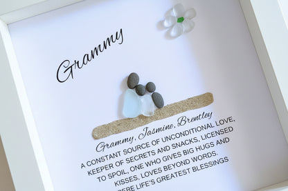 A framed sea glass and pebble art piece with the word 'Grammy' written on it, placed inside a white frame, with a personalized message to a grandmother.