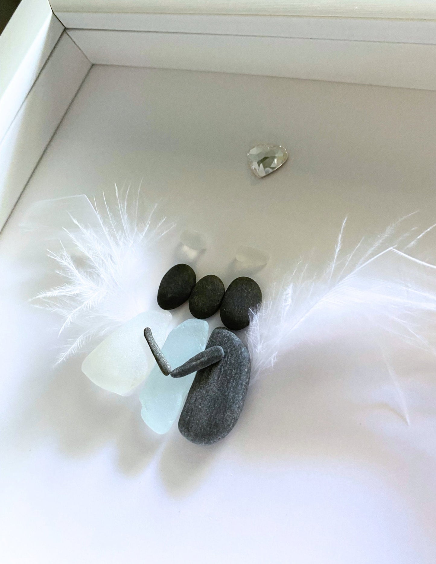 A memorial gift featuring a sea glass art piece with a feather wings hugging a sea glass human, displayed on a white surface.