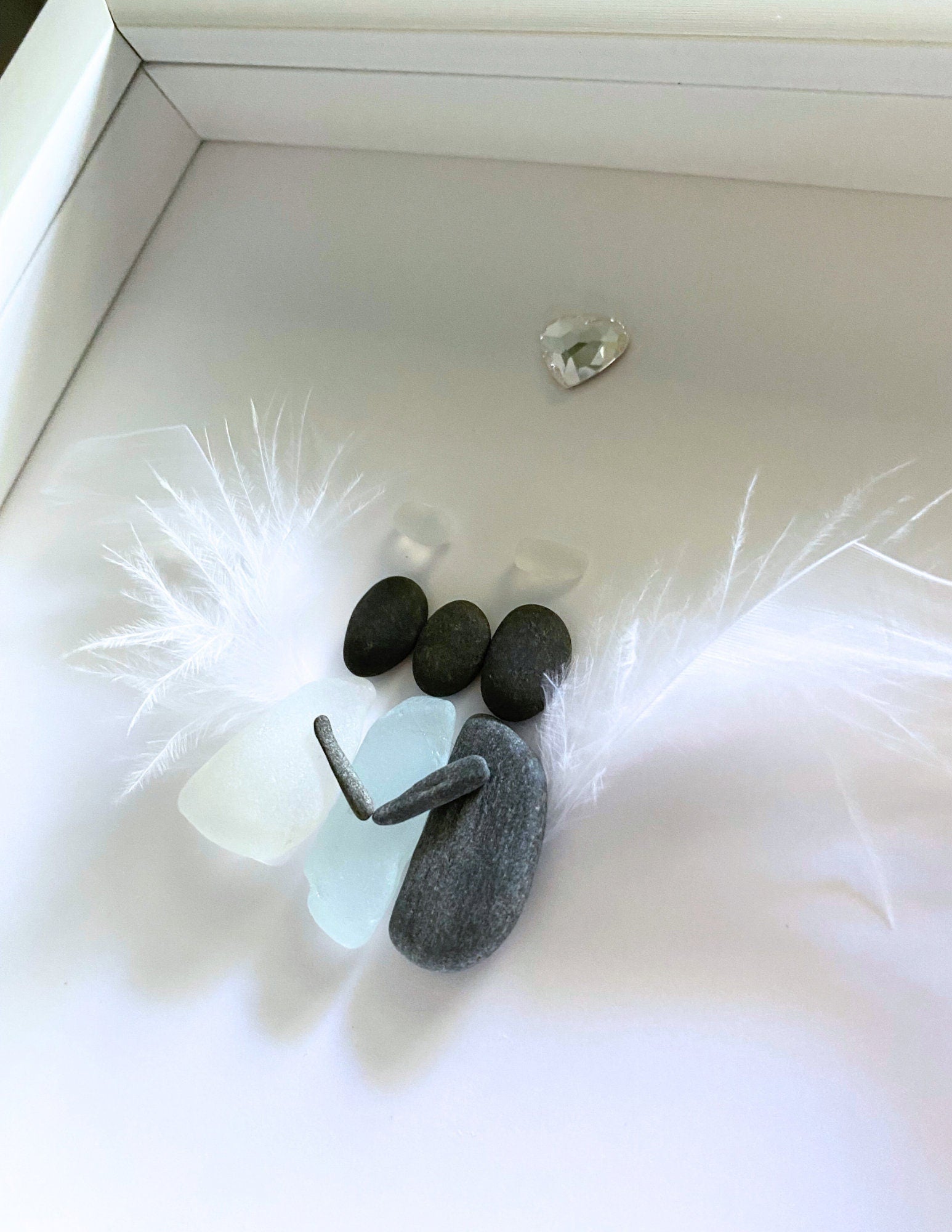 A memorial gift featuring a sea glass art piece with a feather wings hugging a sea glass human, displayed on a white surface.