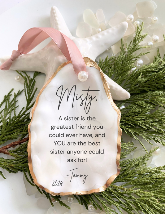 A personalized Christmas ornament oyster shell design, featuring a quote about being a sister, along with greenery and pearls embellishments.