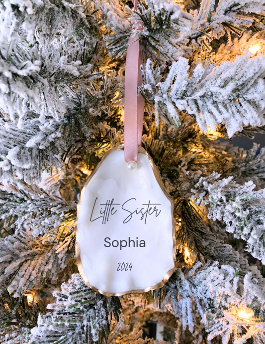 A personalized white oyster shell Christmas ornament with gold gilding and the text 'Little Sister' and a name 'Sophia' along with a date '2020' written on it, hanging on a Christmas tree with a pink ribbon.
