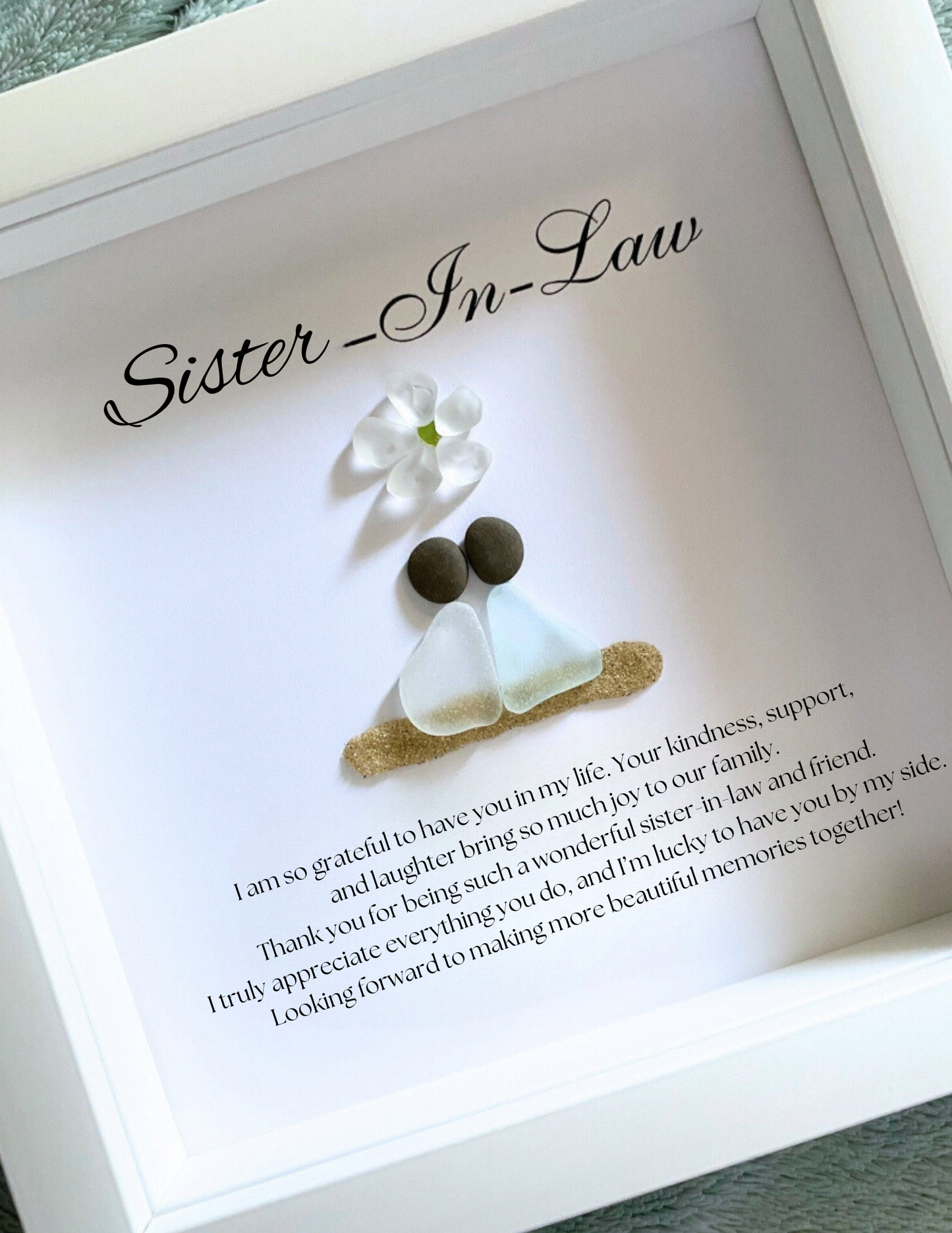 A framed personalized sea glass art piece with the inscription 'Sister-in-Law' on top, a white flower, and two  sea glass figures representing the sisters-in-law. The artwork is enclosed in a white frame.
