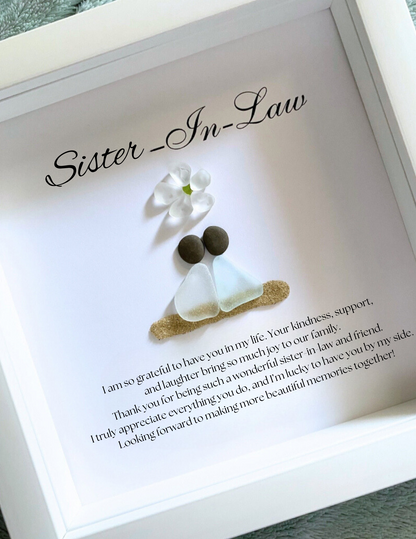 A framed personalized sea glass art piece with the inscription 'Sister-in-Law' on top, a white flower, and two  sea glass figures representing the sisters-in-law. The artwork is enclosed in a white frame.