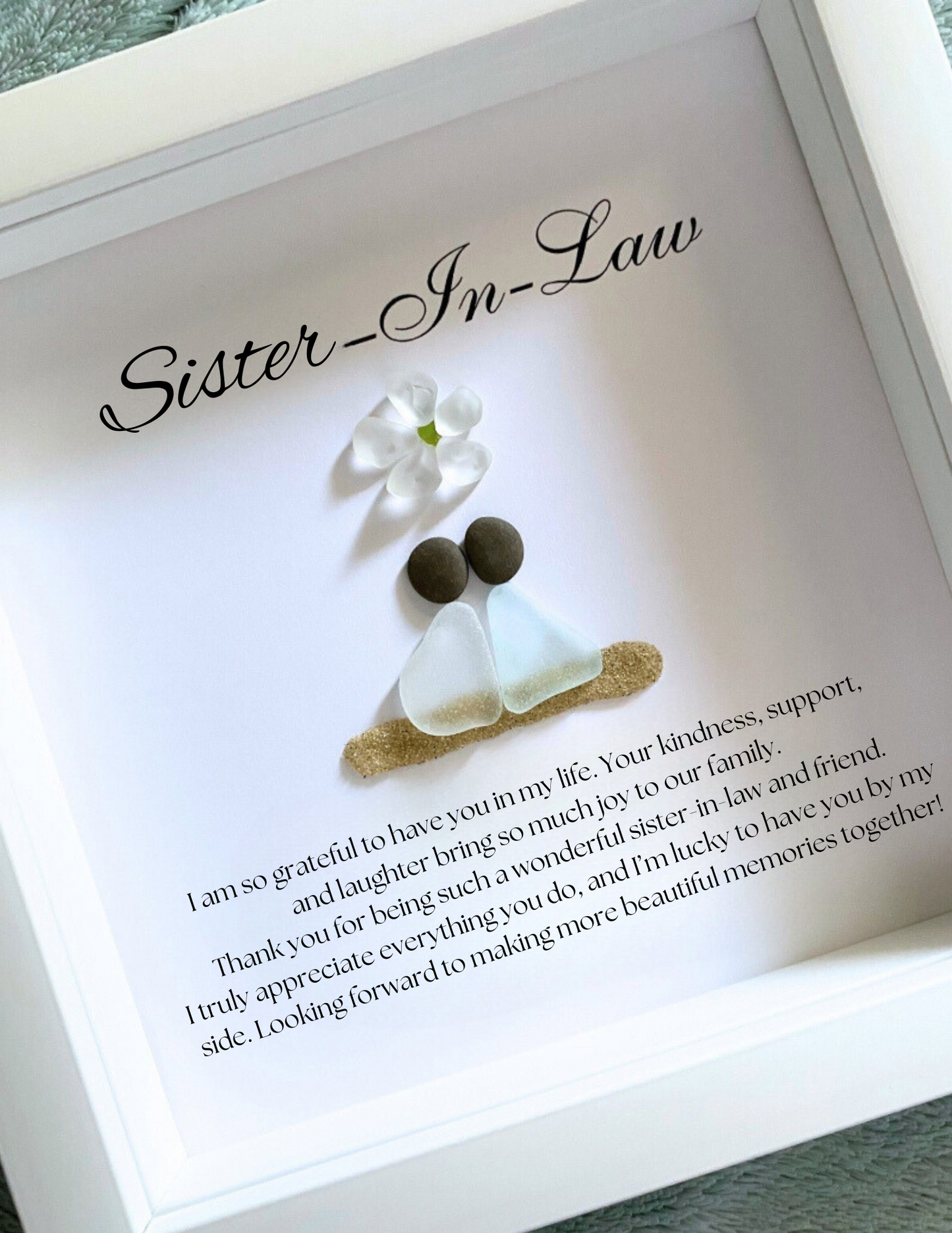 A framed personalized sea glass art piece with the inscription 'Sister-in-Law' on top, a white flower, and two  sea glass figures representing the sisters-in-law. The artwork is enclosed in a white frame.