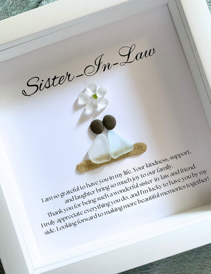 A framed personalized sea glass art piece with the inscription 'Sister-in-Law' on top, a white flower, and two  sea glass figures representing the sisters-in-law. The artwork is enclosed in a white frame.
