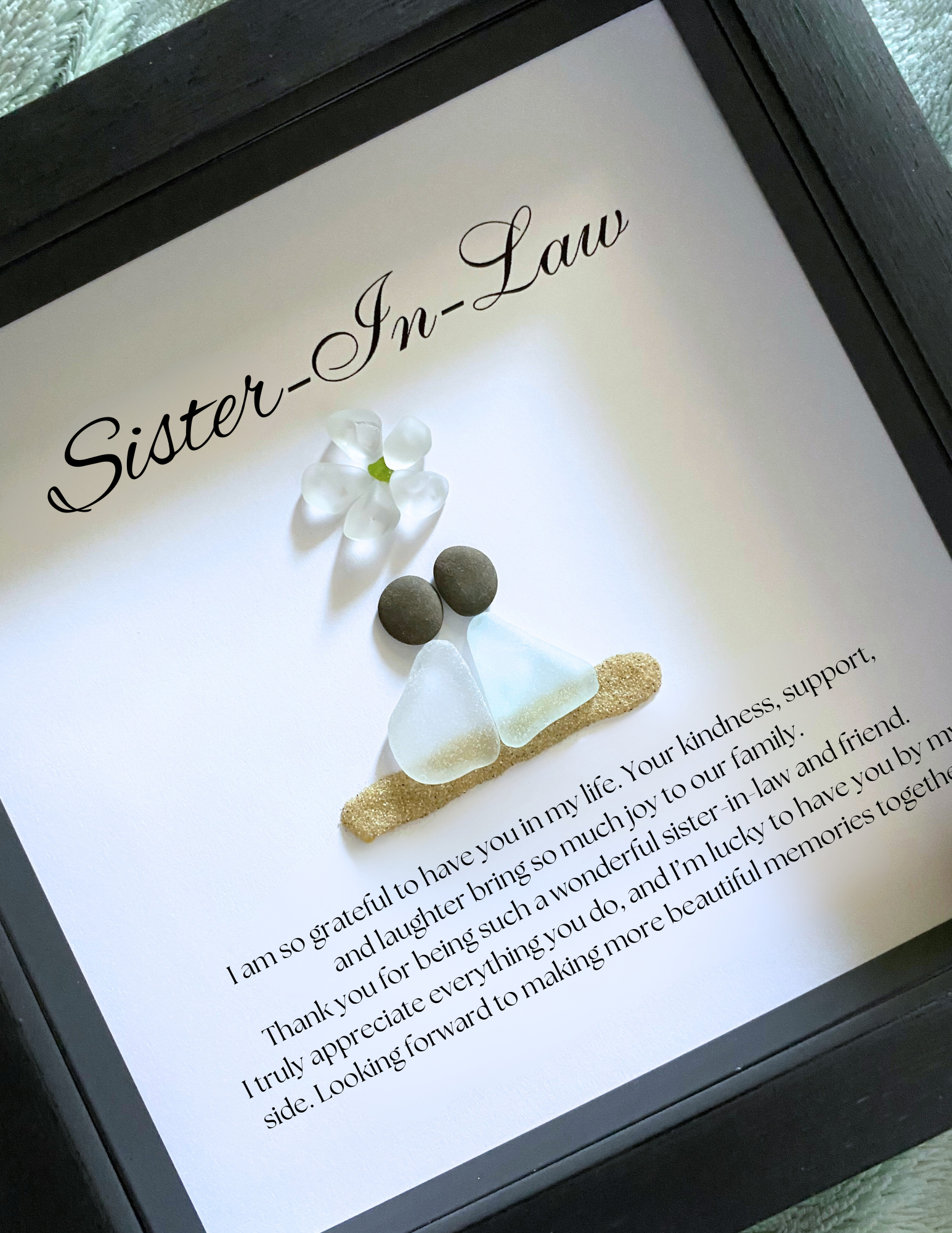 A framed personalized sea glass art piece with the inscription 'Sister-in-Law' on top, a white flower, and two  sea glass figures representing the sisters-in-law. The artwork is enclosed in a black frame.