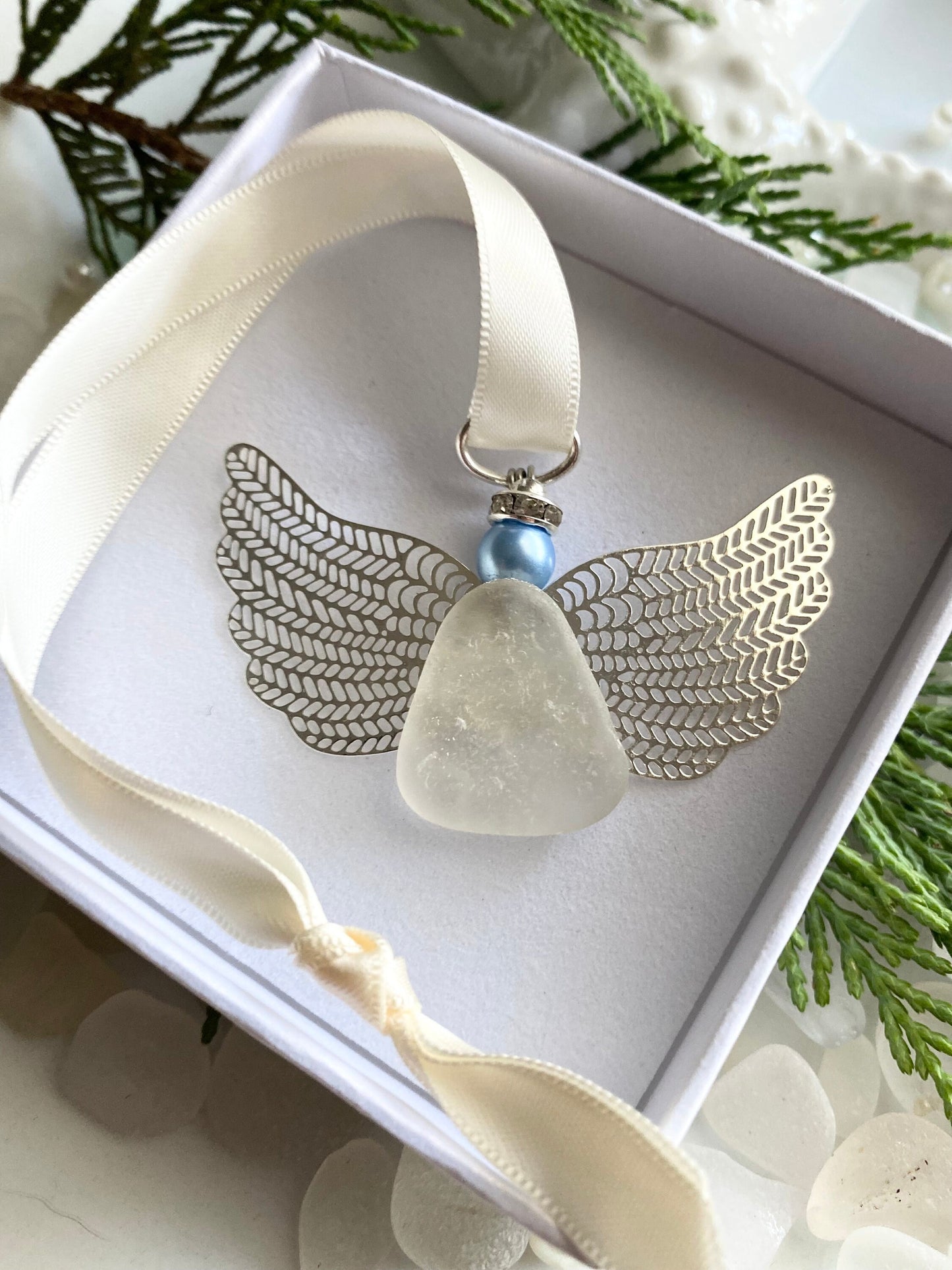 A silver-colored angel-shaped ornament made of sea glass with a blue pearl head, displayed in an open white box with a satin ribbon, set against a natural background with sea glass accents.