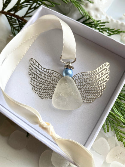 A silver-colored angel-shaped ornament made of sea glass with a blue pearl head, displayed in an open white box with a satin ribbon, set against a natural background with sea glass accents.